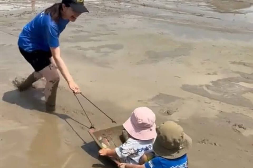 田んぼの中でソリに乗った2人のお子様を、青いTシャツを着た女性が楽しそうに引いている農業体験の風景。