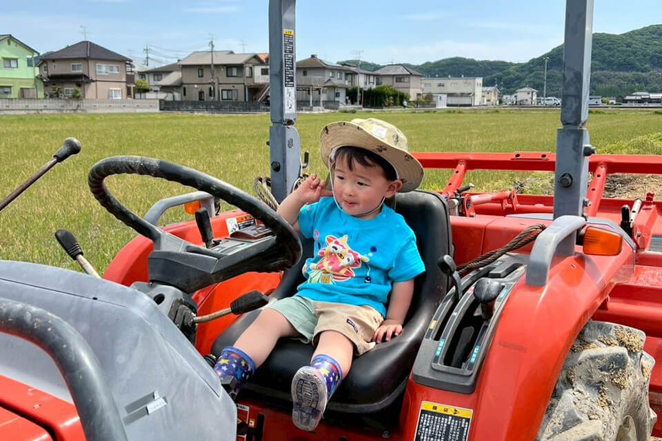 赤いトラクターの運転席に座り、ハンドルを握って得意げな表情を見せる麦わら帽子の男の子。