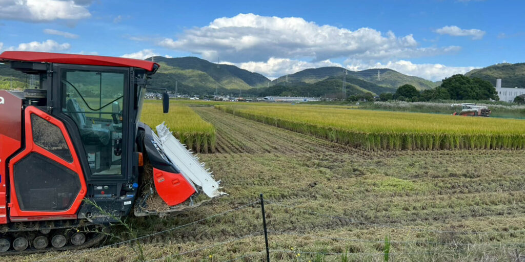 青空と山並みを背景に、効率よく稲刈り作業を行う最新の大型コンバインの側面。